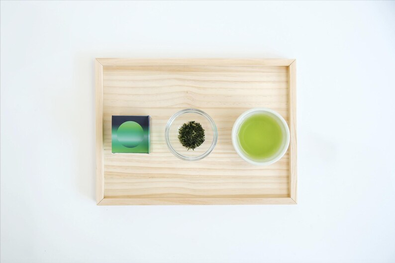 May include: A wooden tray displays a small box with a green gradient design, a small glass bowl of dried green tea leaves, and a white teacup filled with green tea. The items are arranged on a white surface.