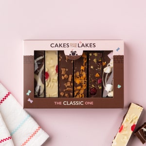 May include: A box of six assorted chocolate bars with the text "Cakes from the Lakes" and "The Classic One" on the front. The bars are arranged in a row and are visible through a clear plastic window. The bars are all different flavours and have different toppings.