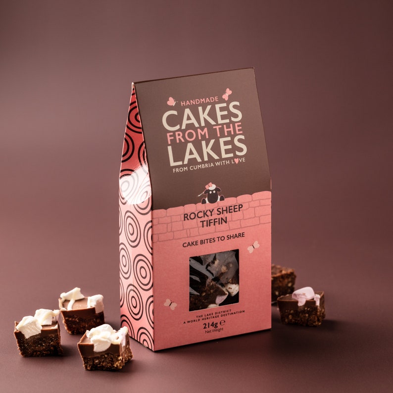 Op de afbeelding: Een roze en bruine doos met Rocky Sheep Tiffin cake hapjes om te delen. De doos heeft een wit schaap met een roze bloem op zijn kop en de tekst "Handmade Cakes From The Lakes From Cumbria With Love". De doos staat op een bruin oppervlak met verschillende cake hapjes eromheen.
