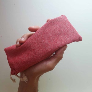 Può includere: Un astuccio rettangolare in tessuto rosso con chiusura a coulisse. L'astuccio è tenuto in una mano, mostrando la sua superficie strutturata e il design semplice. Il colore rosso è uniforme.