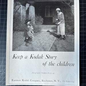 May include: A black and white photograph of a woman taking a picture of a child holding a dog. The text on the image reads "Keep a Kodak Story of the children. Autographic Kodaks £6.50 up. Eastman Kodak Company, Rochester, N. Y., The Kodak City."