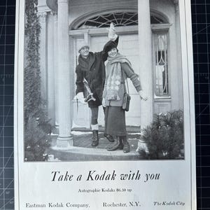 May include: A black and white photograph of two people standing in front of a house. The woman is holding a camera and smiling. The text "Take a Kodak with you" is printed below the image. The text "Autographic Kodaks £6.50 up" is printed below the first line of text. The text "Eastman Kodak Company, Rochester, N.Y. The Kodak City" is printed below the second line of text.