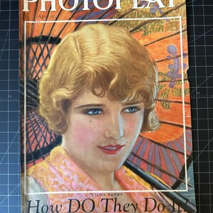 Puede incluir: Portada de la revista Photoplay vintage con un retrato de Vilma Banky. La portada muestra a una mujer con cabello rubio y ojos azules, sobre un fondo de un parasol naranja y morado. El título de la revista y los detalles de la edición son visibles.