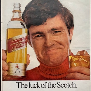 May include: A man in a red turtleneck sweater holds a bottle of Johnnie Walker Red Label Scotch whisky. The bottle is clear glass with a gold label and the text "Johnnie Walker Red Label" is visible. The text "The luck of the Scotch" and "So smooth-world's best selling Scotch" is also visible.