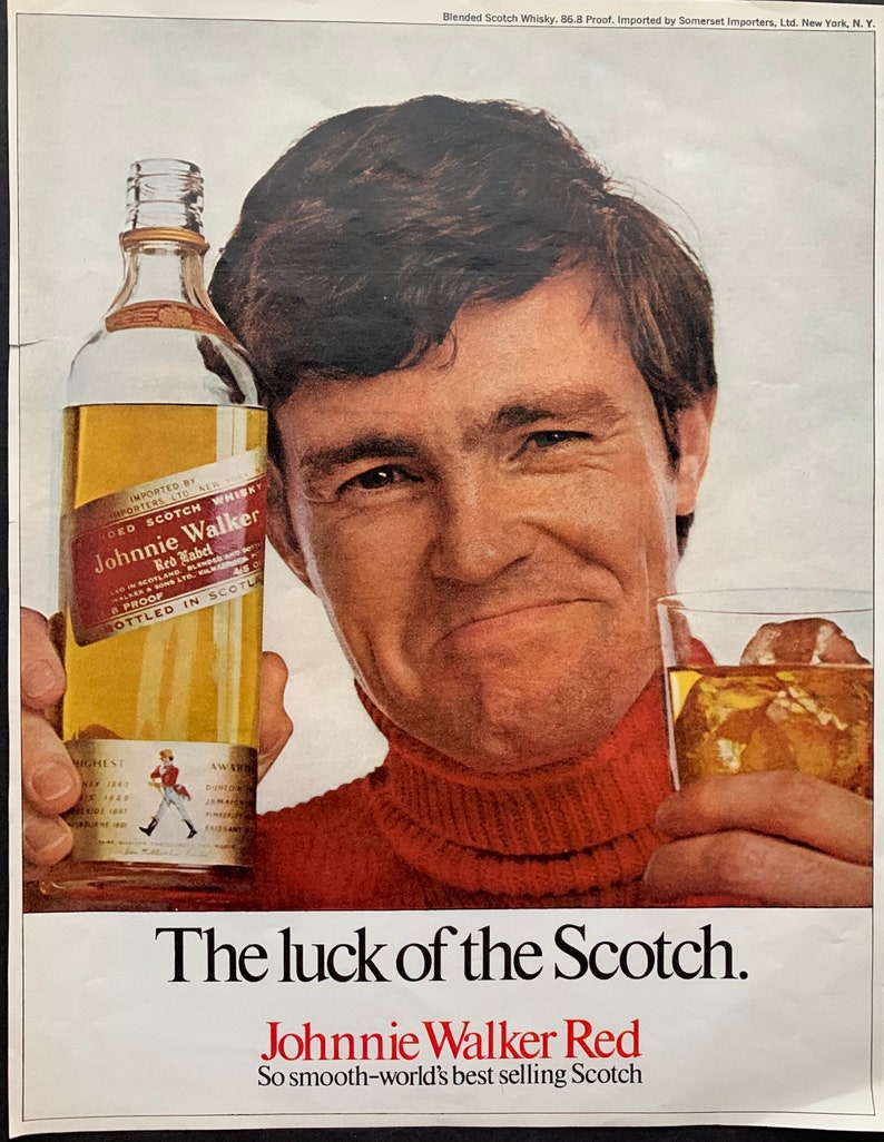 May include: A man in a red turtleneck sweater holds a bottle of Johnnie Walker Red Label Scotch whisky. The bottle is clear glass with a gold label and the text "Johnnie Walker Red Label" is visible. The text "The luck of the Scotch" and "So smooth-world's best selling Scotch" is also visible.