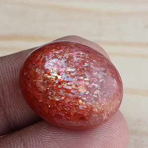 K&ouml;nnte beinhalten: Ein gro&szlig;er, ovaler Sonnenstein-Cabochon in rot-orange mit schimmerndem, irisierendem Effekt. Der Stein hat eine raue, nat&uuml;rliche Textur.