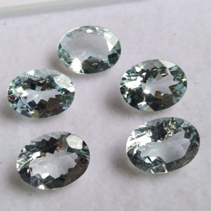 Könnte beinhalten: Fünf ovale Aquamarin-Edelsteine, jeder mit einer hellblauen Farbe und einem facettierten Schliff. Die Edelsteine sind in einem Halbkreis auf einer weißen Oberfläche angeordnet.