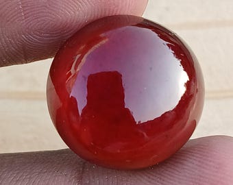 May include: A glossy, deep red marble held between fingers. The spherical stone reflects light, showcasing its smooth, polished surface. The marble's color is a rich, saturated red.
