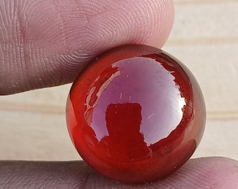 May include: A close-up of a round, glossy, red glass marble. The marble is held between two fingers, showcasing its smooth, reflective surface. The color is a deep, rich red.