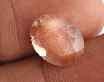 May include: A large, oval-shaped, faceted, sunstone gemstone with a copper-colored sheen. The stone is translucent and has a light pink hue.