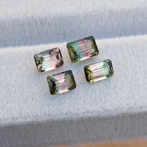 May include: Four rectangular gemstones with a mix of colors, including pink, green, and brown. The gems are set against a white, textured background. The stones have a polished, reflective surface.