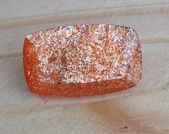 May include: A rectangular, orange sunstone gemstone with a shimmering, iridescent finish. The stone has a rough, textured surface and is set against a light brown wooden background.