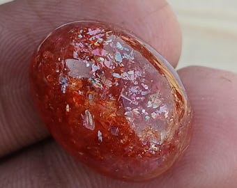 May include: A large, oval-shaped, sunstone cabochon with a red, orange, and gold colour. The stone has a shimmering, iridescent effect.