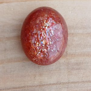 K&ouml;nnte beinhalten: Ein runder, roter Sunstone-Cabochon mit irisierendem Schimmer. Der Stein hat eine glatte, polierte Oberfl&auml;che und liegt auf einem hellbraunen Holzuntergrund.
