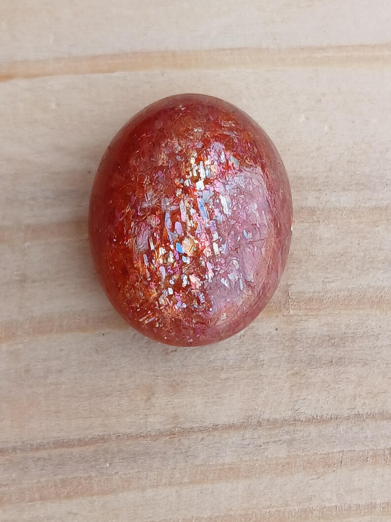 K&ouml;nnte beinhalten: Ein runder, roter Sunstone-Cabochon mit irisierendem Schimmer. Der Stein hat eine glatte, polierte Oberfl&auml;che und liegt auf einem hellbraunen Holzuntergrund.