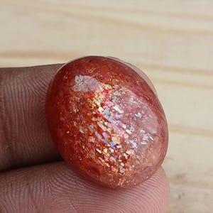 K&ouml;nnte beinhalten: Ein gro&szlig;er, ovaler, roter Sunstone-Cabochon mit irisierendem Schimmer. Der Stein hat eine glatte, polierte Oberfl&auml;che und liegt auf einem h&ouml;lzernen Hintergrund.