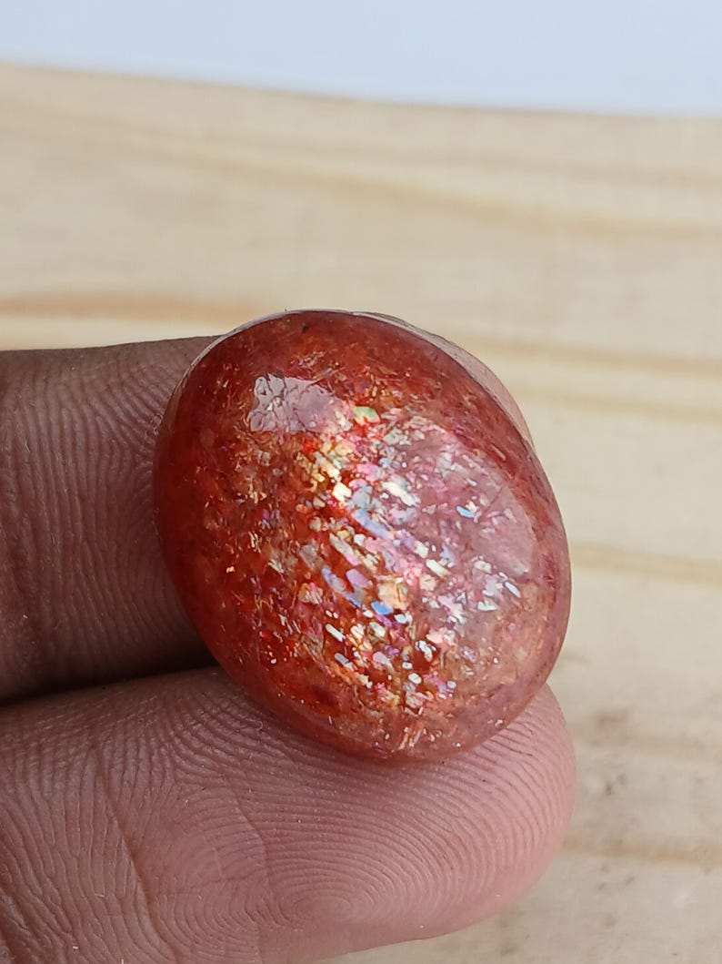K&ouml;nnte beinhalten: Ein gro&szlig;er, ovaler, roter Sunstone-Cabochon mit irisierendem Schimmer. Der Stein hat eine glatte, polierte Oberfl&auml;che und liegt auf einem h&ouml;lzernen Hintergrund.