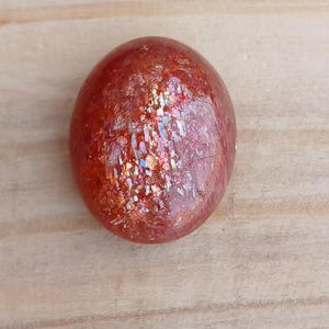K&ouml;nnte beinhalten: Ein runder, roter Sunstone-Cabochon mit irisierendem Schimmer und einer glatten, polierten Oberfl&auml;che.