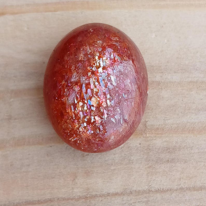 K&ouml;nnte beinhalten: Ein runder, roter Sunstone-Cabochon mit irisierendem Schimmer und einer glatten, polierten Oberfl&auml;che.