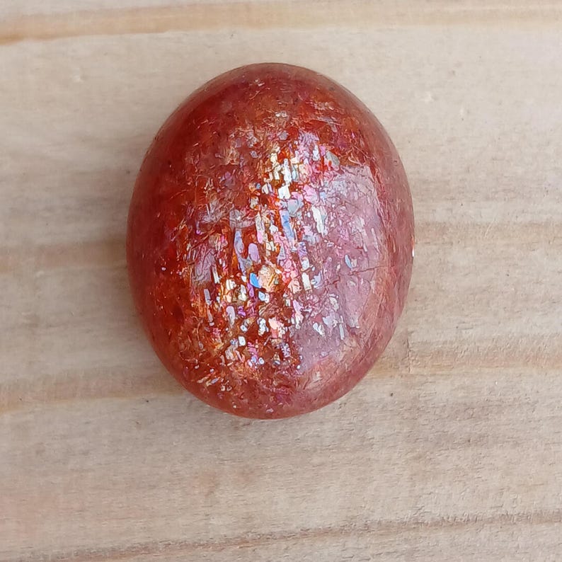 K&ouml;nnte beinhalten: Ein runder, roter Sunstone-Cabochon mit irisierendem Schimmer. Der Stein hat eine glatte, polierte Oberfl&auml;che und liegt auf einem hellbraunen Holzuntergrund.
