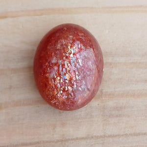 K&ouml;nnte beinhalten: Ein runder, polierter Sunstone-Cabochon in rot-orange mit irisierendem Schimmer.