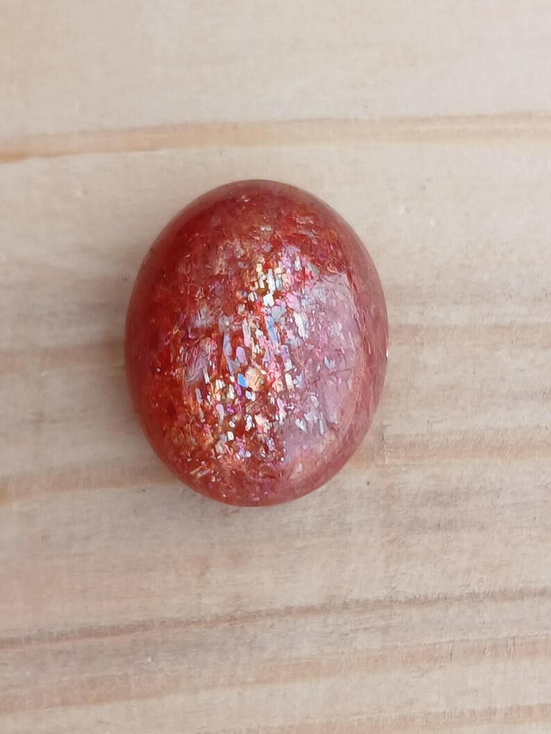 K&ouml;nnte beinhalten: Ein runder, polierter Sunstone-Cabochon in rot-orange mit irisierendem Schimmer.