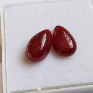 Puede incluir: Dos piedras preciosas rojas en forma de pera, probablemente rubíes, se muestran sobre una superficie blanca. Las piedras preciosas son lisas y pulidas, reflejando la luz.