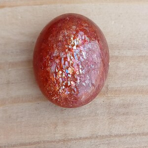 K&ouml;nnte beinhalten: Ein runder, polierter Sunstone mit einer rotbraunen Farbe und einem irisierenden Glanz.
