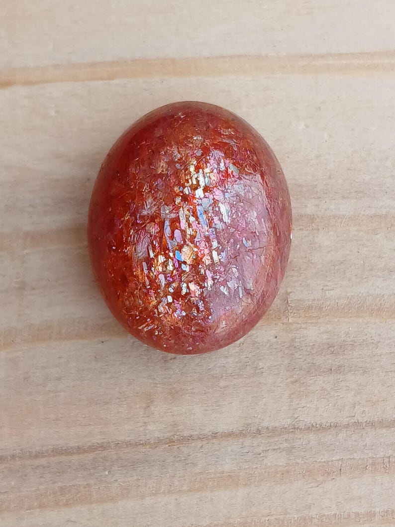K&ouml;nnte beinhalten: Ein runder, polierter Sunstone mit einer rotbraunen Farbe und einem irisierenden Glanz.