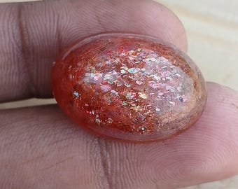 May include: A red sunstone cabochon with a shimmering, iridescent finish. The stone is oval-shaped and has a smooth, polished surface.
