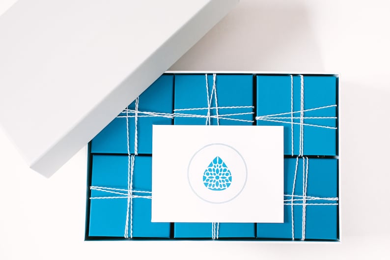 May include: A white box with a blue and white logo on the lid is open, revealing a set of 12 small blue boxes tied together with white string.