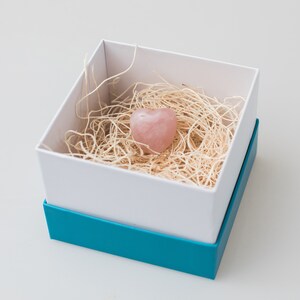 May include: A pink heart-shaped crystal is nestled in a white box filled with shredded paper. The box has a teal blue lid.