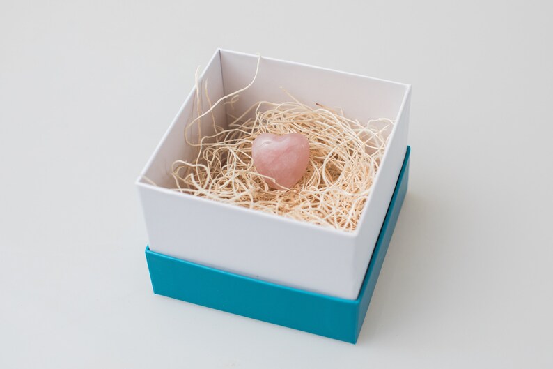 May include: A pink heart-shaped crystal is nestled in a white box filled with shredded paper. The box has a teal blue lid.