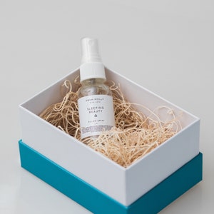 May include: A small white bottle with a spray nozzle, labeled "Sleeping Beauty Pillow Spray" in a white box with a teal blue lid. The bottle is sitting on a bed of shredded paper.