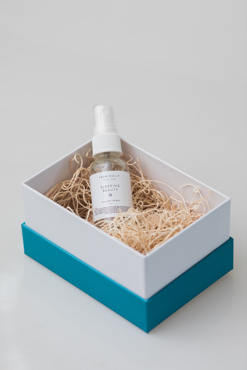 May include: A small white bottle with a spray nozzle, labeled "Sleeping Beauty Pillow Spray" in a white box with a teal blue lid. The bottle is sitting on a bed of shredded paper.