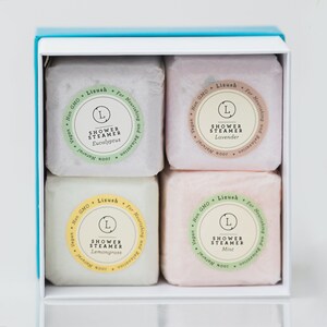 May include: A white box with a light blue lid contains four shower steamers. The steamers are in a grid pattern and are labeled with the scents: Eucalyptus, Lavender, Lemongrass, and Mint. Each steamer has a green and white label with the text "Shower Steamer" and the scent name.