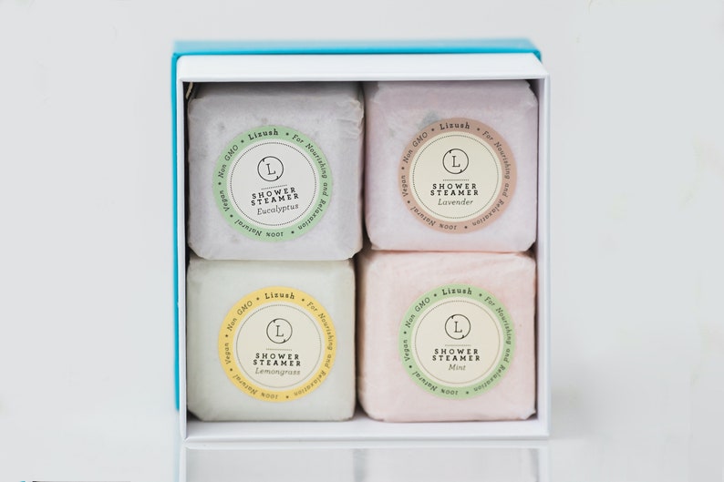 May include: A white box with a light blue lid contains four shower steamers. The steamers are in a grid pattern and are labeled with the scents: Eucalyptus, Lavender, Lemongrass, and Mint. Each steamer has a green and white label with the text "Shower Steamer" and the scent name.