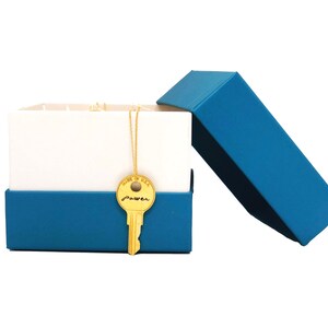 May include: A blue gift box with a white lid is open, revealing a gold key necklace with the word "power" inscribed on the key. The necklace is on a white background.