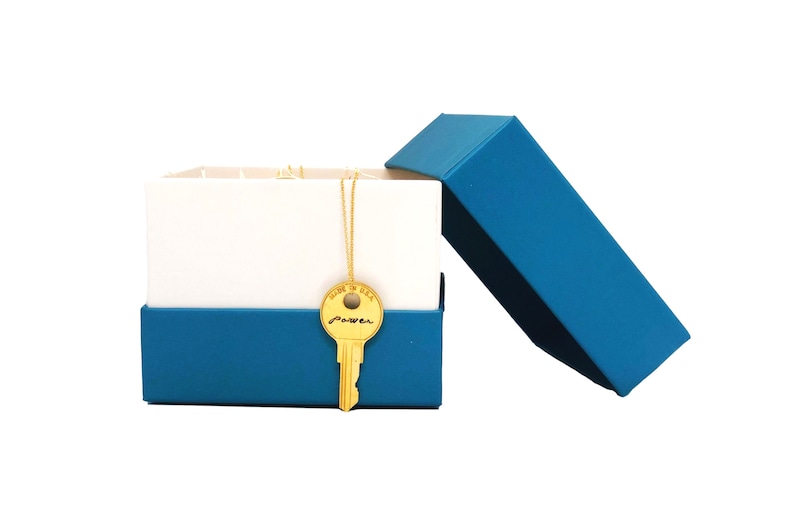 May include: A blue gift box with a white lid is open, revealing a gold key necklace with the word "power" inscribed on the key. The necklace is on a white background.