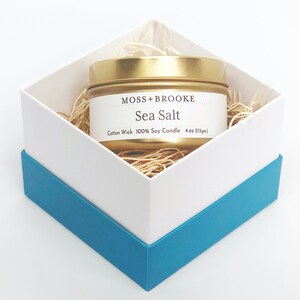 May include: A gold tin candle with a white label that reads "Moss + Brooke Sea Salt Cotton Wick 100% Soy Candle 4 oz (113g)". The candle is in a white box with a blue trim.