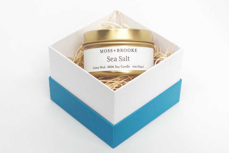 May include: A gold tin candle with a white label that reads "Moss + Brooke Sea Salt Cotton Wick 100% Soy Candle 4 oz (113g)". The candle is in a white box with a blue trim.
