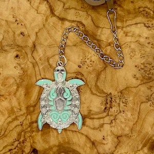 以下が含まれることがあります： チェーンとターコイズ色の亀のチャームが付いたシルバーのティーインフューザー。インフューザーはメッシュデザインの球体です。亀のチャームは、渦巻きや線で詳細なデザインが施されています。