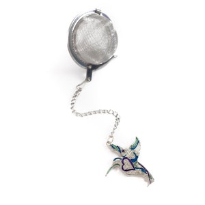 May include: Silver tea infuser with a chain and a hummingbird charm. The hummingbird is decorated with blue, green, and purple enamel.