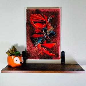 May include: A rectangular wall art piece featuring a comic book character in a red cape and blue suit with silver chains, set against a dark red and black speckled background. Beside it sits an orange fox-shaped planter with a small green succulent.