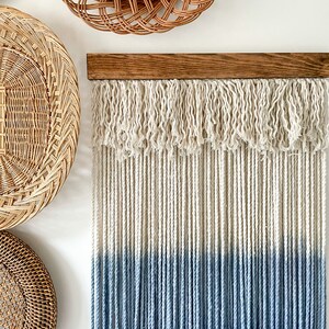 May include: A macrame wall hanging with a wooden dowel at the top. The macrame is made of white and blue cotton cord. The cord is tied in a fringe pattern at the top and hangs in straight strands. The bottom half of the macrame is dyed a light blue color.