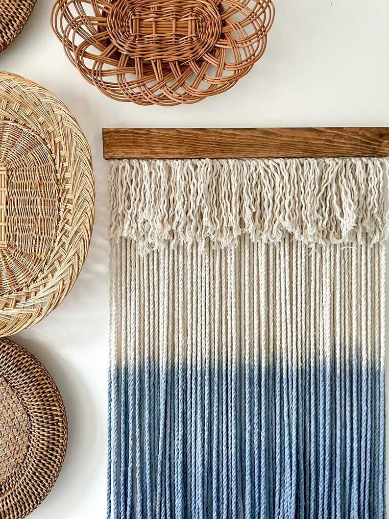 May include: A macrame wall hanging with a wooden dowel at the top. The macrame is made of white and blue cotton cord. The cord is tied in a fringe pattern at the top and hangs in straight strands. The bottom half of the macrame is dyed a light blue color.