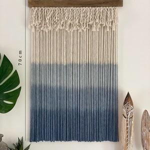 May include: A blue and white ombre macrame wall hanging with a wooden dowel at the top. The hanging measures 46 cm wide and 70 cm long.