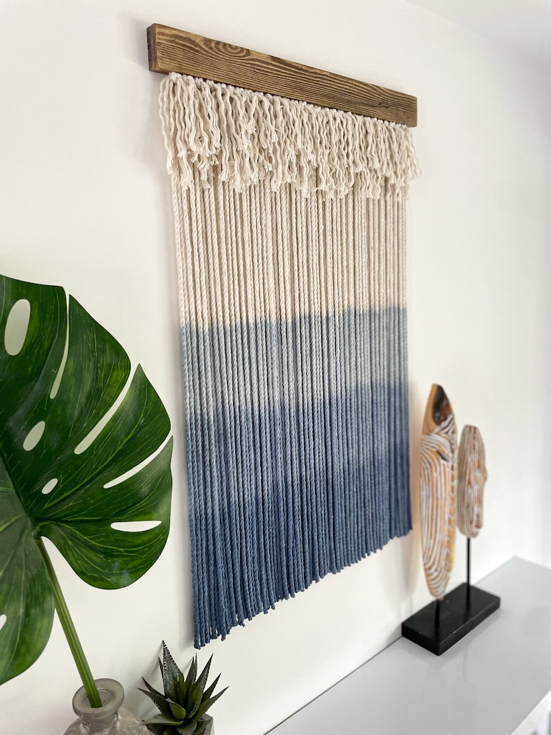 May include: A blue and white ombre macrame wall hanging with a wooden dowel at the top. The macrame is made with cotton cord and has a fringe at the bottom.