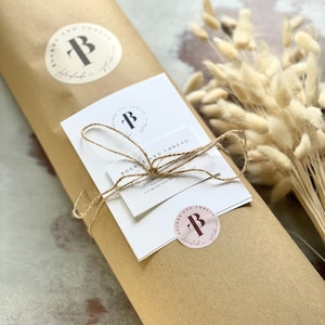 May include: A brown paper package wrapped with twine and a white card with the text "Bourne and Thread" and a logo with the letter "B". The package is decorated with two round stickers with the same logo.