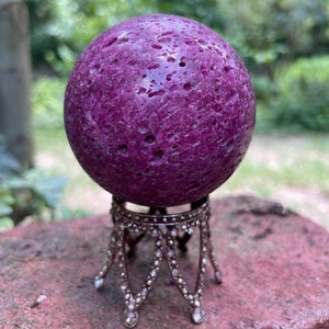 May include: A deep purple, round, polished gemstone sphere on a decorative metal stand with a beaded design. The sphere has a rough, textured surface.